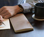 Person writing in a notebook with a black mug on a table