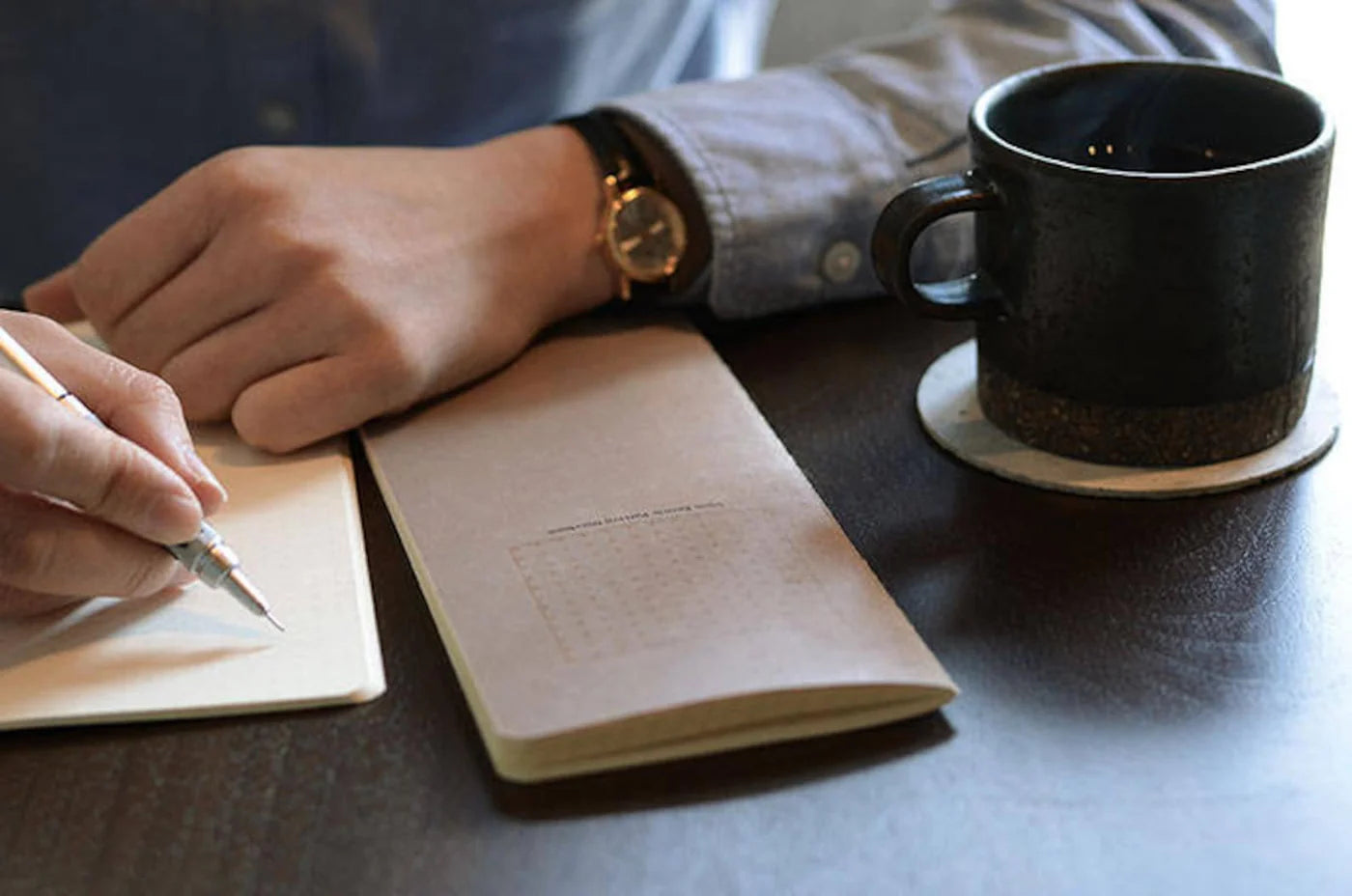 Person writing in a notebook with a black mug on a table