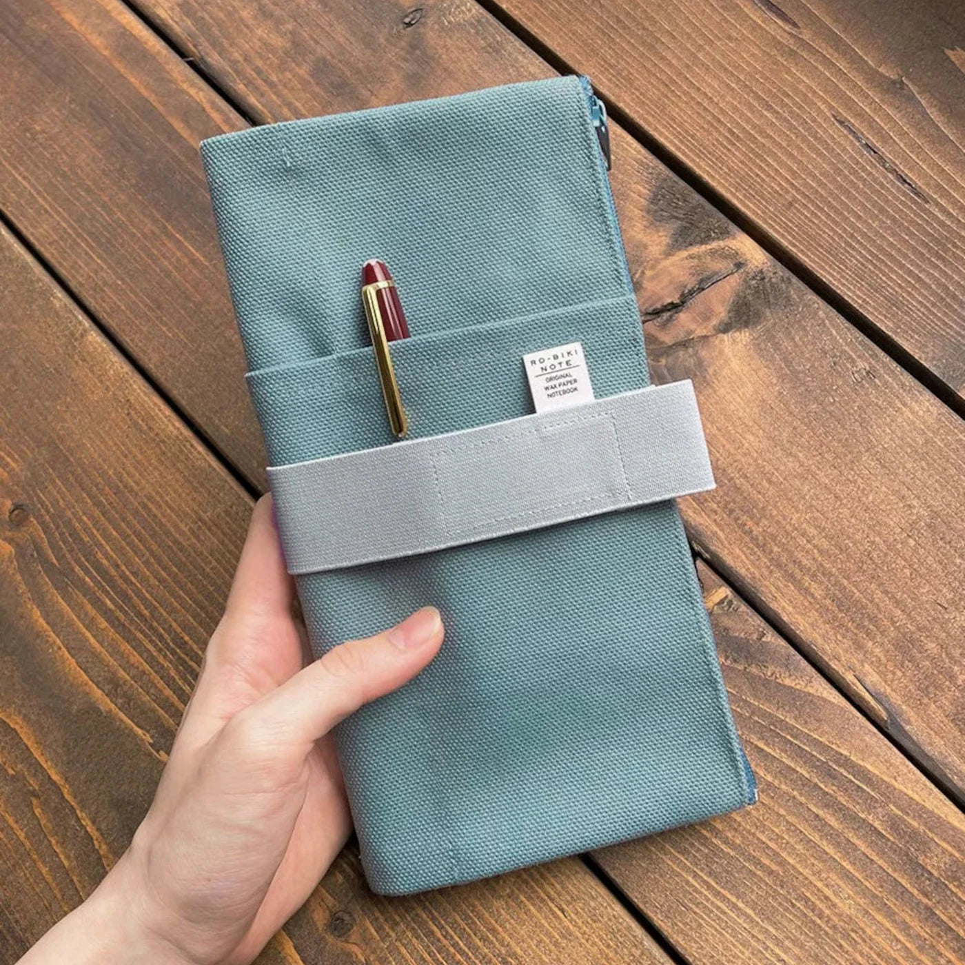 Teal fabric wallet with a pen held by a hand on a wooden surface