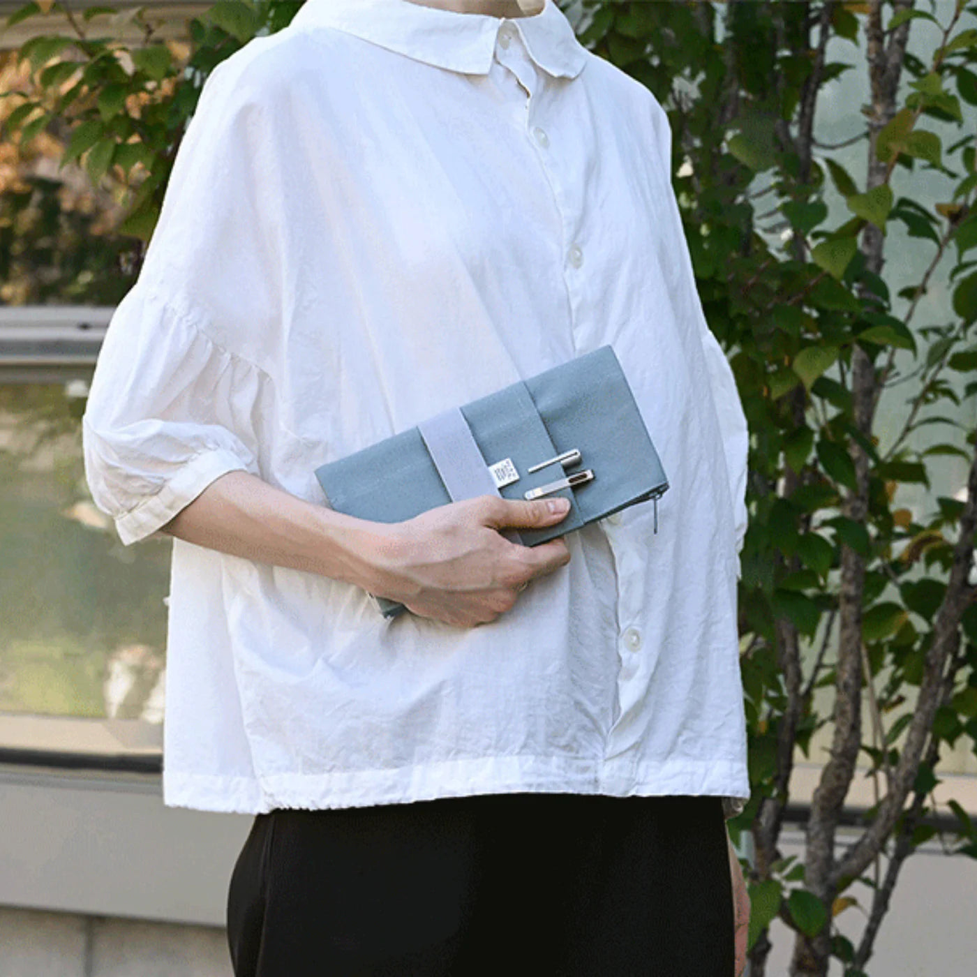 Person wearing a white shirt holding a gray clutch bag outdoors.
