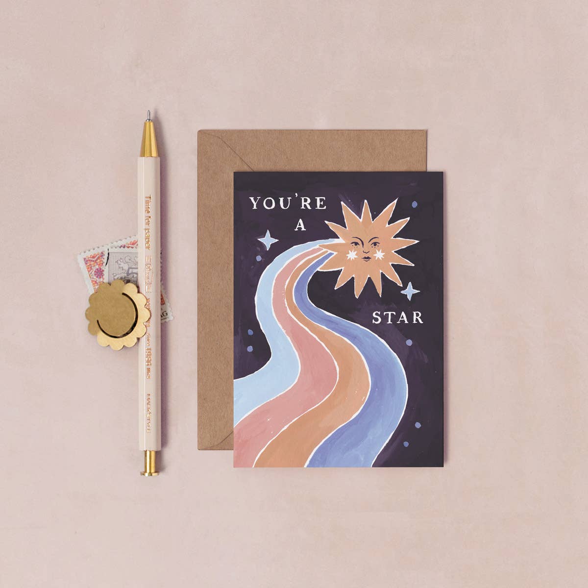 Greeting card with sun and moon illustration and 'You're a star' text, next to a pen and envelope on a beige background.
