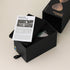 Black box containing a bicolor sandglass with an open lid and product information sheet.