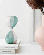 Green and pink hourglass on a stack of books with a pink vase and dried plants in the background.