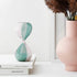 Green and pink hourglass on a stack of books with a pink vase and dried plants in the background.