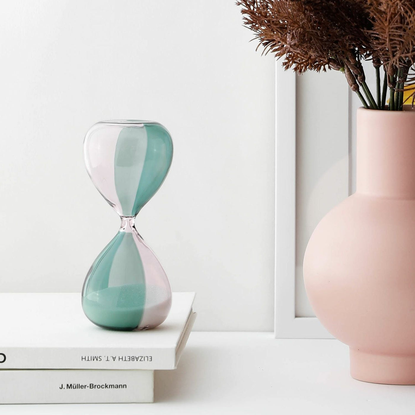 Green and pink hourglass on a stack of books with a pink vase and dried plants in the background.
