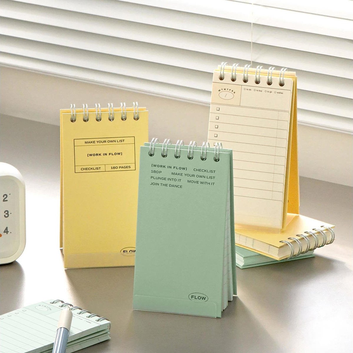 Three spiral-bound flip agenda notebooks in pastel yellow, mint green, and cream are arranged on a tabletop in front of window blinds. The notebooks have lined pages and checklist sections printed in black.