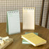 Two spiral-bound notepad books and a matching yellow hardcover notebook arranged on a wooden table, with a checkered curtain and wooden chair visible in the background.