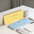A yellow spiral-bound desk calendar and a light blue horizontal notepad labeled FLOW are arranged on a white desk with a keyboard, mouse, phone, pencil, and a small white device.