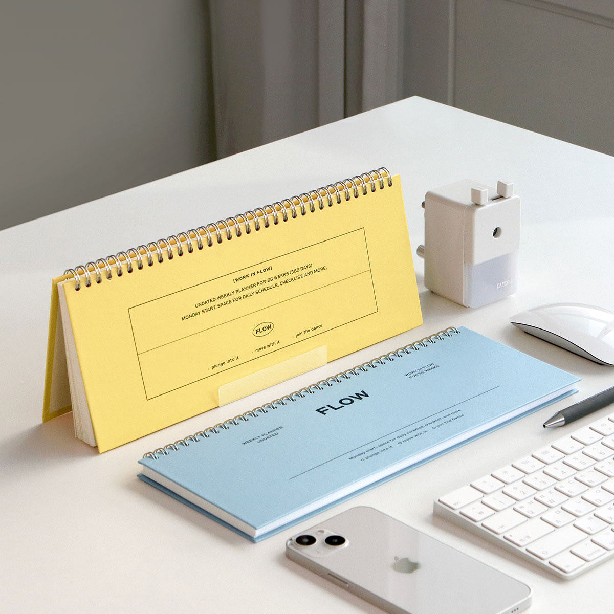 A yellow spiral-bound desk calendar and a light blue horizontal notepad labeled FLOW are arranged on a white desk with a keyboard, mouse, phone, pencil, and a small white device.