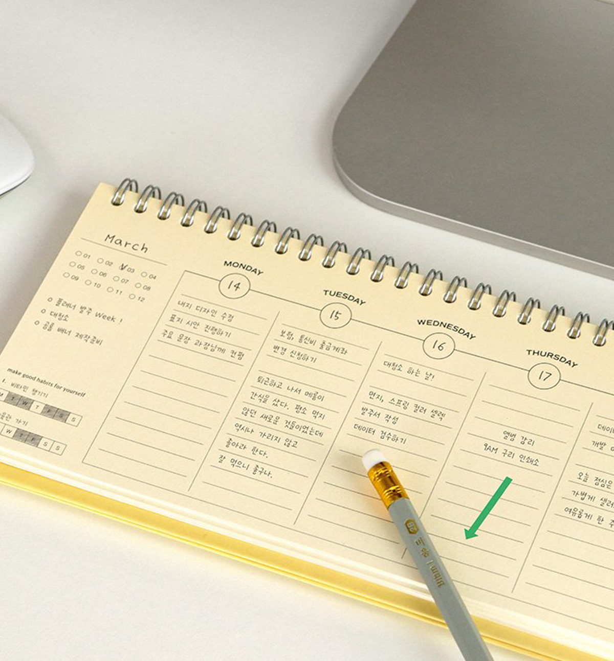 A spiral-bound planner page showing a weekly schedule layout in English and Korean, photographed on a light desk with part of a laptop visible and a gray pen resting on the page.