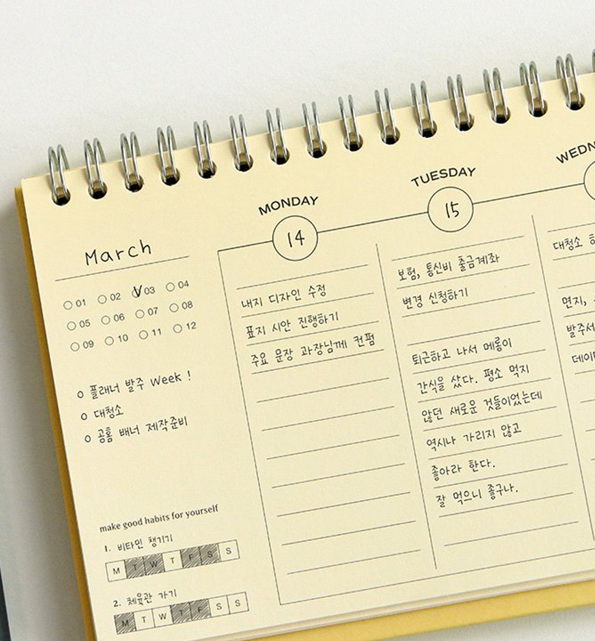 A spiral-bound planner page shown close up, with cream paper, silver metal spiral binding, and printed sections for Monday and Tuesday with handwritten notes in Korean.