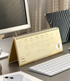 A gold spiral-bound desk calendar standing open on a gray desk, surrounded by a computer monitor, keyboard, tablet, and phone in a styled office setting.