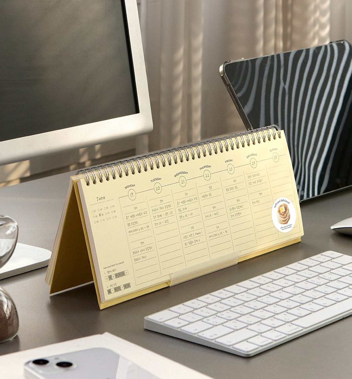 A gold spiral-bound desk calendar standing open on a gray desk, surrounded by a computer monitor, keyboard, tablet, and phone in a styled office setting.