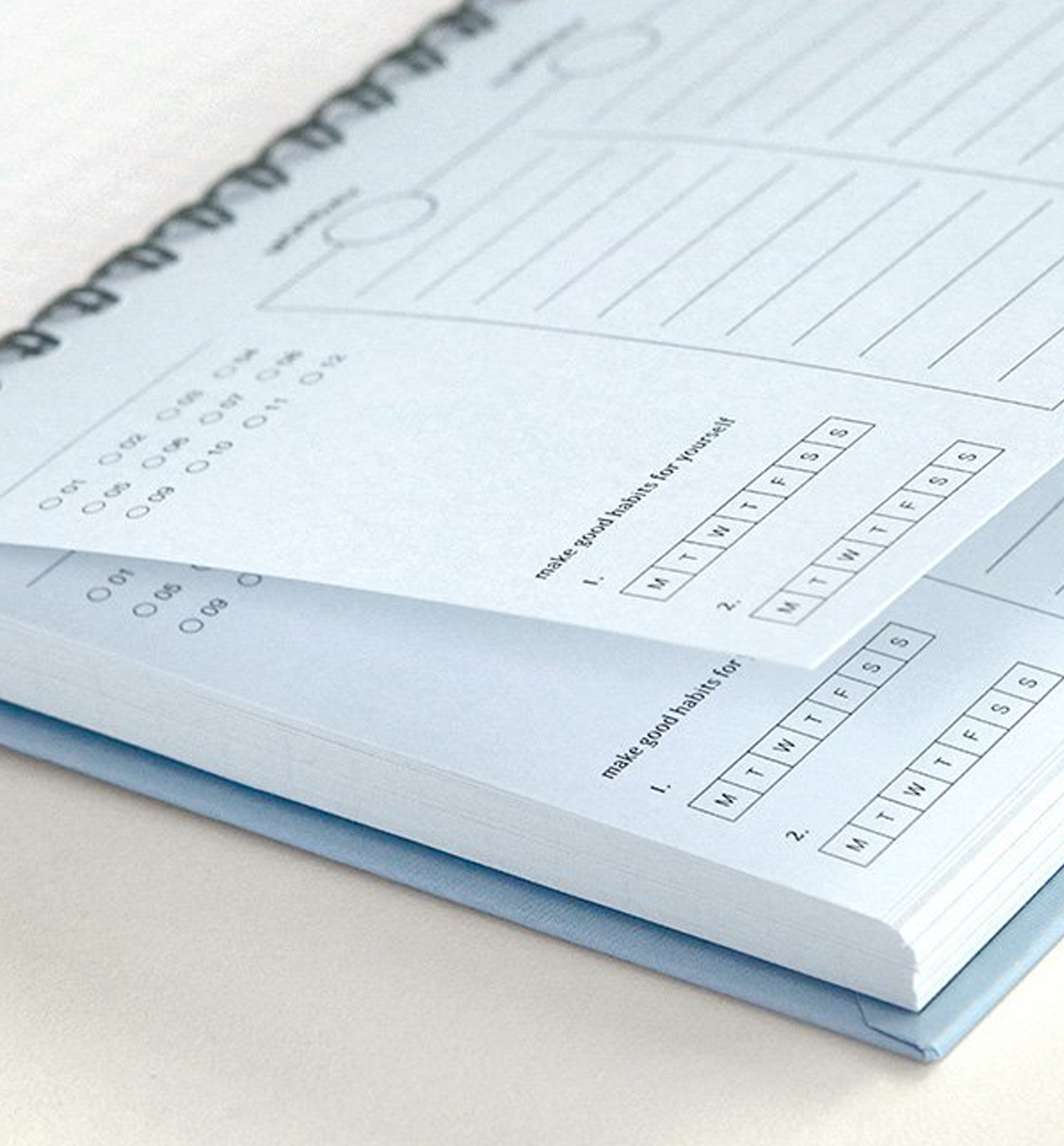 A close-up of a spiral-bound planner or notebook showing light blue lined pages with printed daily planning boxes and a calendar section, resting on a light neutral surface.
