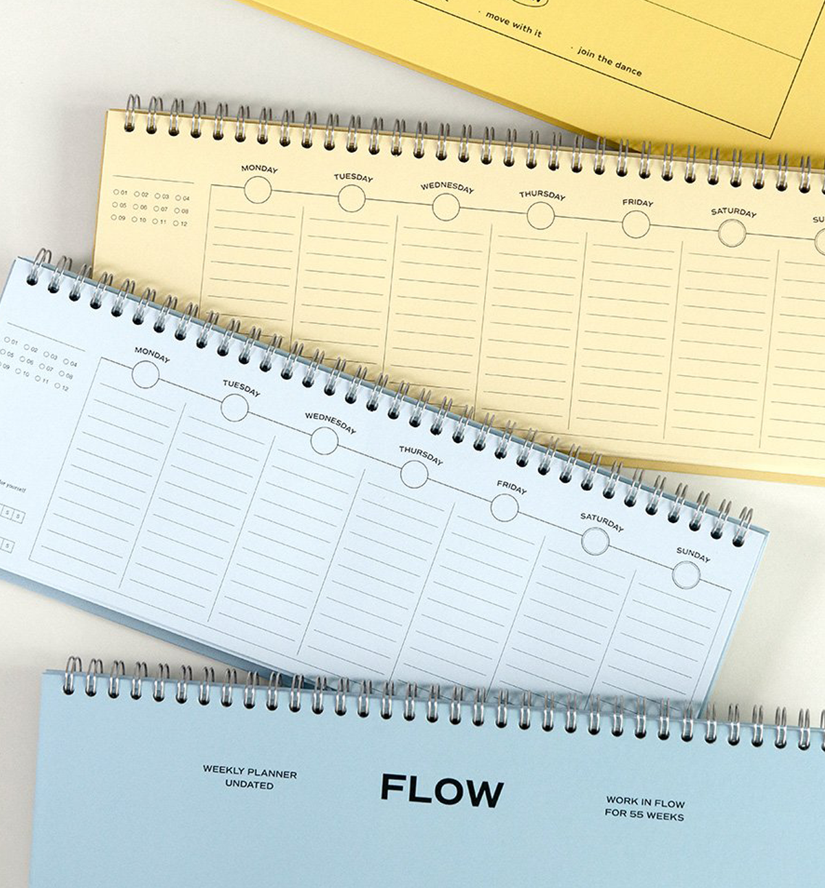 Three spiral-bound weekly planner notebooks in pastel blue, cream, and yellow, arranged diagonally on a white surface. The bottom notebook features the brand name 'FLOW' printed prominently on its cover.