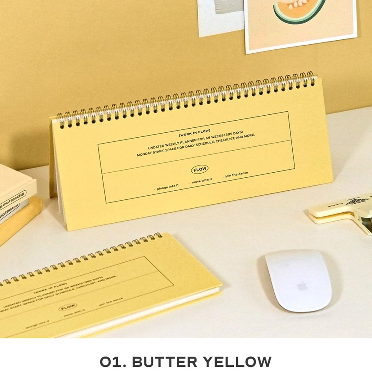 A butter yellow spiral-bound desk calendar and matching notepad displayed on a light beige desk with a white computer mouse, a gold pen, and a small stack of books. The text 'O1.