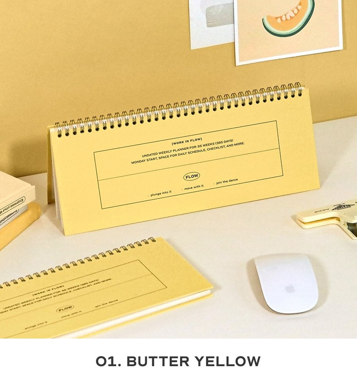 A butter yellow spiral-bound desk calendar and matching notepad displayed on a light beige desk with a white computer mouse, a gold pen, and a small stack of books. The text 'O1.