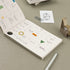 Open planner with colorful icons on a beige surface with a pen and paper clip.