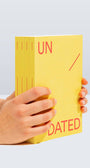 Yellow book titled 'UNDATED' held by a hand on a light gray background
