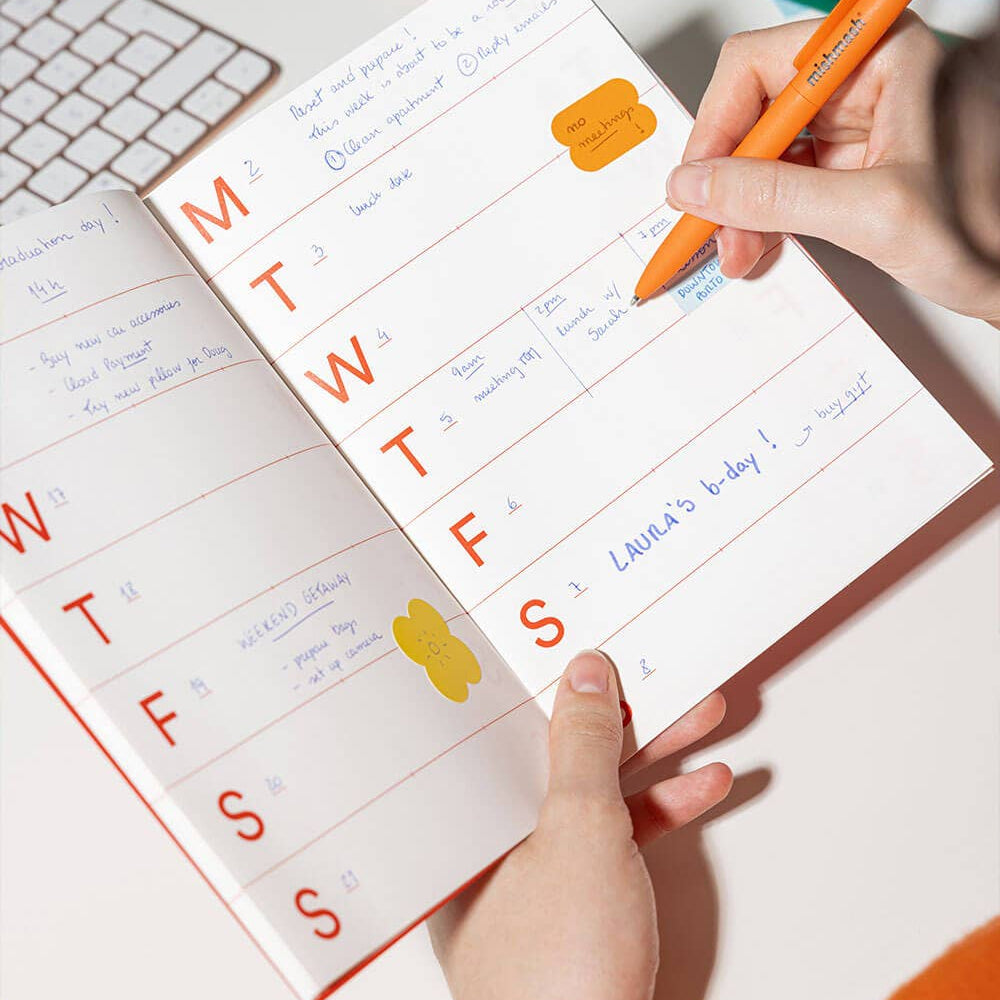 Person using a planner with stickers and an orange pen on a white surface.