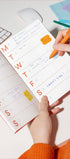 Person using a planner with stickers and an orange pen on a white surface.