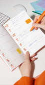 Person using a planner with stickers and an orange pen on a white surface.