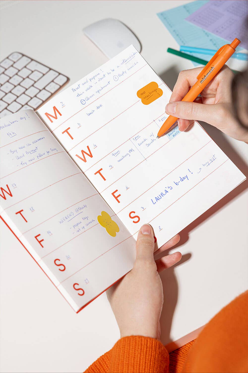 Person using a planner with stickers and an orange pen on a white surface.